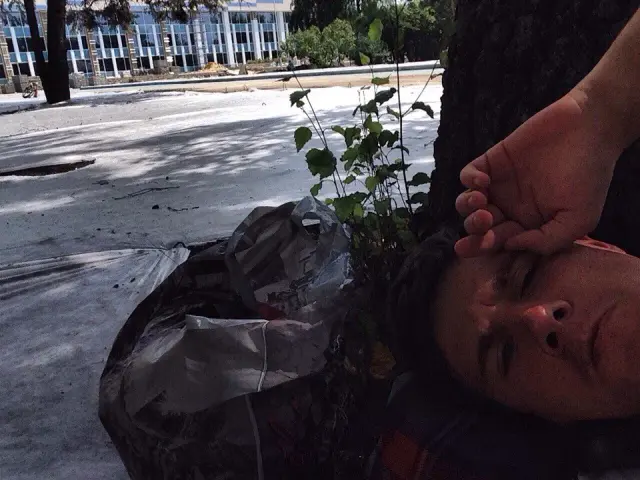 Alexey lying beside a small plant and bags on a sidewalk